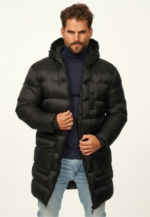Black padded long coat with a hood, featuring a zippered front, four large pockets, and a textured finish. Worn over a dark turtleneck.