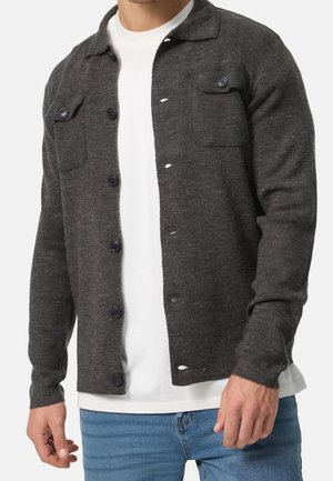 Man draagt een donkergrijze, gebreide cardigan met knopen over een wit T-shirt en een blauwe spijkerbroek, zichtbaar van nek tot heupen.