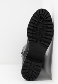 Semelle de botte noire avec un motif de crampons profond et marquage de taille 37, positionnée à la verticale contre une surface blanche de coin.