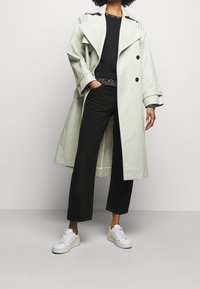 Persona vistiendo un abrigo trench beige claro, suéter negro, pantalones negros cortos y zapatillas blancas, de pie con una mano en el bolsillo.