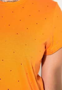 Orange kortärmad T-shirt gjord av ett mjukt tyg, prydd med små, runda, reflekterande detaljer som är jämnt fördelade över framsidan.
