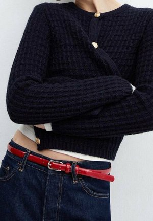 Person iført en tekstureret sort sweater med gyldne knapper, mørkeblå jeans og et skinnende rødt bælte med en sølvspænde.