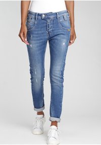 Onderlichaam van een persoon die blauwe distressed opgerolde skinny jeans en witte veterschoenen draagt tegen een effen achtergrond.