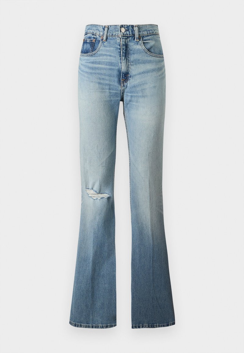 Jean en denim bleu clair taille haute avec légère décoloration, détail usé au genou gauche et jambes évasées.