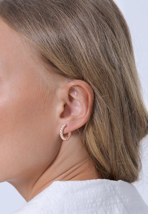 Rosaguld hoopsmykke med små klare ædelsten, der har en glat finish og cirkulær form, båret i en models øre.