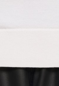 Weißer gerippter Sweaterbund mit strukturiertem Finish, über einer glatten schwarzen Lederschicht, die kontrastierende Materialien und klare Linien präsentiert.