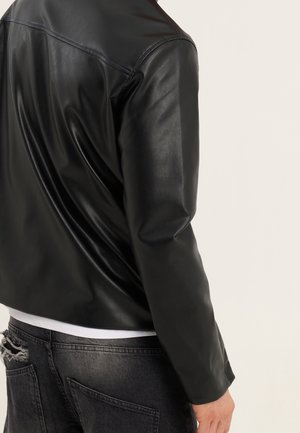 Giacca di pelle nera con un design elegante e una consistenza liscia. Presenta un colletto rotondo e una silhouette corta. Indossata sopra una camicia bianca.