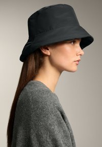 Pălărie neagră tip bucket, cu boruri late, textură netedă și un design structurat. Pur și simplu cu un pulover gri tricotat. Vedere din profil lateral.