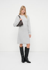Donna in un vestito a maglia grigia fino al ginocchio, stivali neri alti fino al ginocchio, che tiene una borsa marrone scuro a tracolla, in piedi contro uno sfondo bianco.