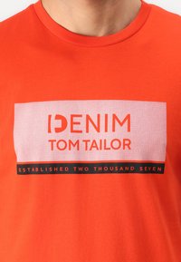 Oranžinė medvilninė marškinė su grafikiniu printu, kuriame užrašyta "DENIM TOM TAILOR" ir "ESTABLISH TWO THOUSAND SEVEN" juoda ir balta spalvomis.