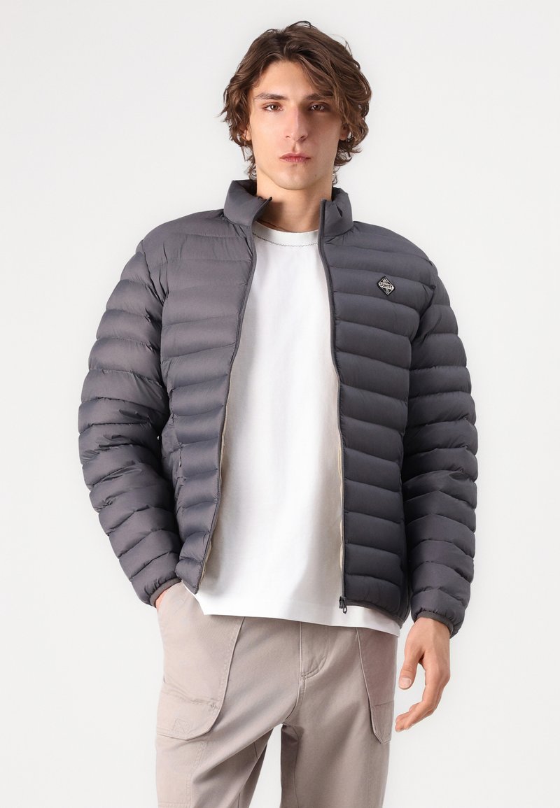 Doudoune grise, design matelassé, col montant, fermeture éclair sur le devant, tissu léger. Portée par-dessus une chemise blanche, avec un pantalon de couleur neutre.