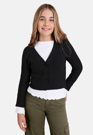 Schwarze gerippte Strickjacke mit V-Ausschnitt und Knopfverschluss, getragen über einem weißen Rüschenshirt. Das Modell ist mit olivgrünen Hosen gestylt.