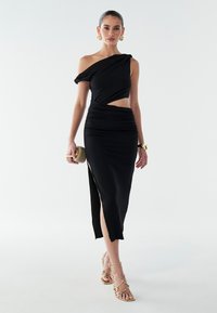 Musta ühekohaline kleit volditud detailide ja külgsuuna lõhiku jaoks. Sile kangas ja keha ligi istuv siluett. Kuldsed rihmadega kontsakingad ja käekott.