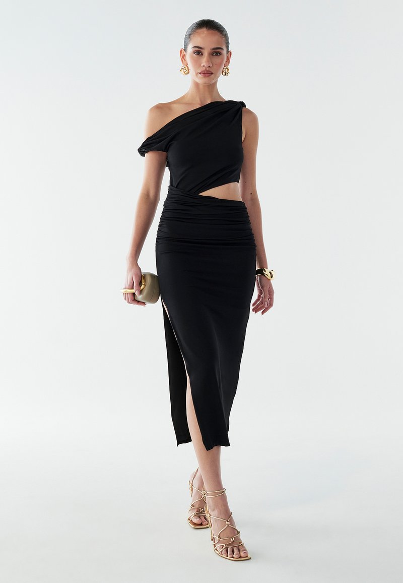 Musta ühekohaline kleit volditud detailide ja külgsuuna lõhiku jaoks. Sile kangas ja keha ligi istuv siluett. Kuldsed rihmadega kontsakingad ja käekott.