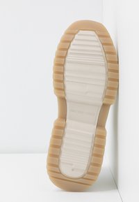 Semelle d’une chaussure beige clair avec des motifs de bande de roulement texturés et un bord extérieur en gomme rainuré, placée verticalement contre un fond blanc en angle.