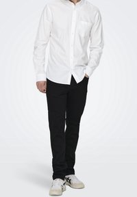 Man in een witte button-up shirt, zwarte broek en witte sneakers staat met één hand in zijn zak tegen een effen achtergrond.