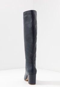 Botte noire en cuir jusqu'au genou avec un bout carré, un talon bloc, une texture lisse et des détails de couture le long de l'arrière.