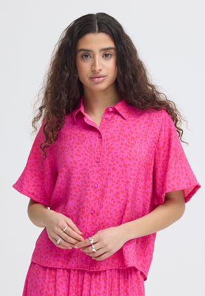 Jeune femme aux longs cheveux bouclés portant une chemise à manches courtes boutonnée à motifs roses et une jupe assortie, les mains jointes devant elle.