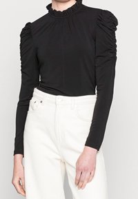 Persona che indossa una maglia nera a maniche lunghe con spalle arricciate e colletto arricciato, abbinata a pantaloni bianchi a vita alta.