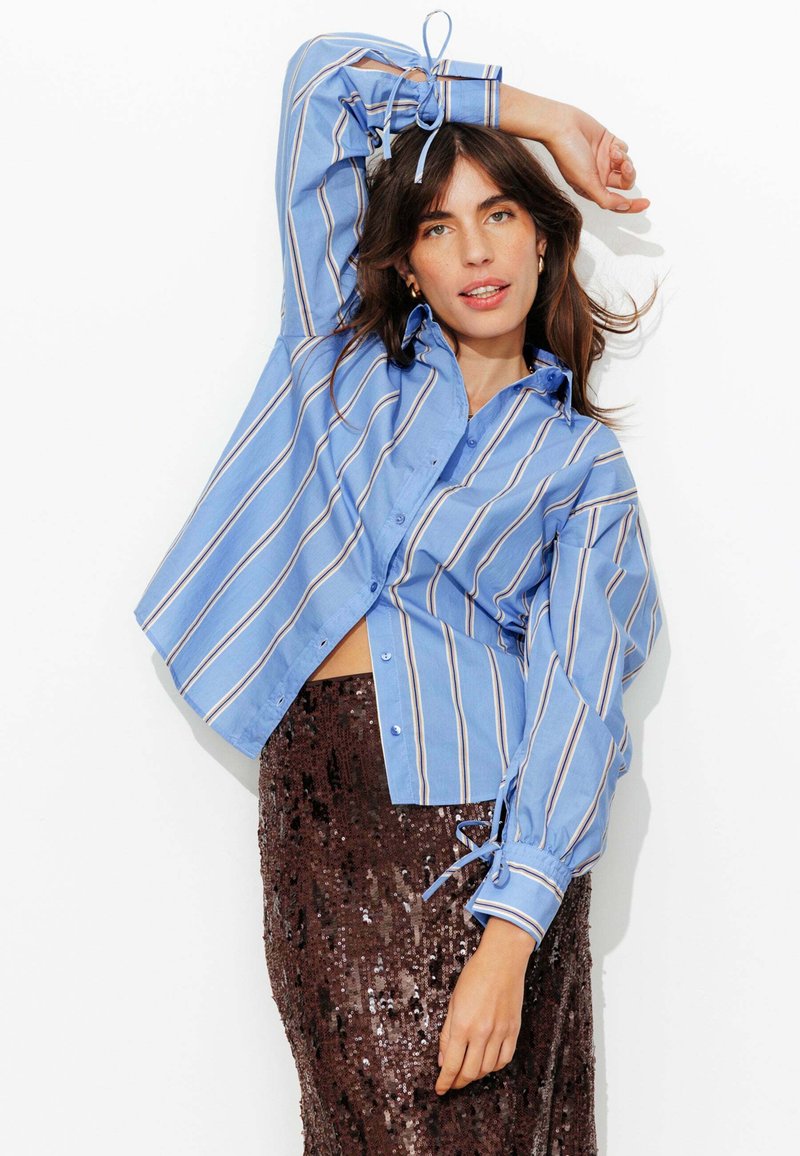Chemise boutonnée bleu à rayures avec manches longues, détails de nouage aux poignets et coupe décontractée, associée à une jupe marron à sequins.