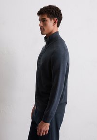 Dunkelblaues Zip-up Sweatshirt mit hohem Kragen, strukturiertem Muster, gerippten Bündchen und Saum. Das Modell steht seitlich vor einem schlichten Hintergrund.