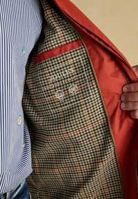 Raudono džinsinio švarko viduje yra dėmėtas raštas šviesiai rusvos ir juodos spalvos, su prekės ženklu ir priežiūros simboliais. Išorinis audinys atrodo lygus.