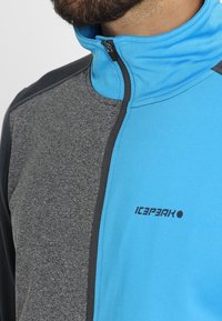 Chaqueta con cremallera que presenta una mezcla de colores azul, gris y negro. La tela es suave, con un diseño ajustado y un logo en el pecho.