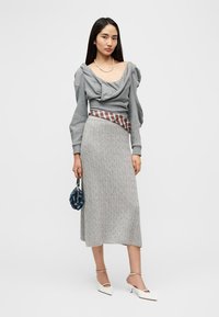 Γκρι πουλόβερ με λαιμόκοψη τύπου κουκούλας, υφασμάτινη φούστα ανοιχτού γκρι με υφή πλεξίματος, καρό ζώνη, μπλε τσάντα και λευκά ψηλοτάκουνα παπούτσια με μυτερή μύτη.