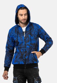 Blaue Kapuzenjacke mit zippbarer Front, ausgestattet mit grafischen schwarz-weißen Mustern von Tieren und tribalem Design. Schwarze Bündchen und Saum betonen den lebhaften Look.
