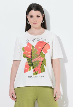 Frau mit glattem dunklem Haar trägt ein weißes T-Shirt mit grünem und korallenfarbenem Blätterdruck und dem Schriftzug „Great Things Modern View“ sowie grüne Hosen.