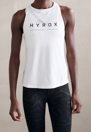 Atletyczna osoba ubrana w biały bezrękawnik z logo "HYROX" i czarne legginsy w wzory, stojąca na jednolitym tle.