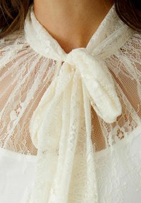 Kant blouse met een hoge kraag en een zachte, geknoopte sluiting. De stof is ivoorkleurig en toont ingewikkelde kantpatronen en texturen.