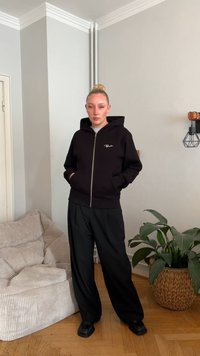 Casaco preto com fecho de correr à frente e bolso frontal, combinado com calças pretas largas e sapatos pretos. Texturas suaves e corte descontraído.