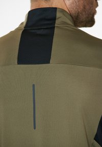 Olivgrüne Sportjacke mit schwarzer Mesh-Besatz auf dem oberen Rücken, mit glatter Oberfläche und einem vertikalen reflektierenden Akzent.