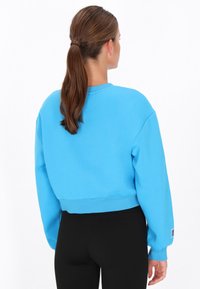 Przycięta niebieska bluza z miękkiego materiału z ściągaczami na rękawach i u dołu. Na lewym rękawie znajduje się metka. Noszona z czarnymi legginsami.