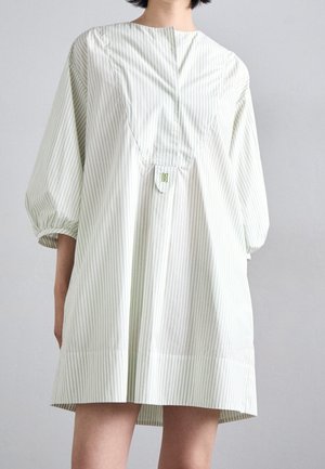 Femme portant une robe ample rayée de couleur claire avec des manches bouffantes trois-quarts et un petit détail de patte sur le devant, sur un fond uni.