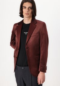 Blazer de terciopelo burdeos con solapas de muesca, cierre frontal con un solo botón y bolsillo. Llevado sobre una camiseta negra y pantalones grises.