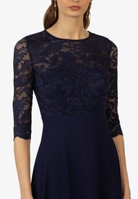 Abito blu navy con corpetto in pizzo e motivi floreali, maniche a tre quarti, che si presenta con una gonna liscia e uniforme.
