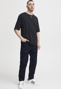 Črna široka majica, temno modre kavbojke, bele superge s črnimi črtami, stoječi položaj, kratki lasje, minimalistični nakit, nevtralna ozadje.