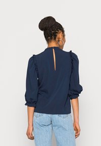 Blusa blu navy con maniche a sbuffo e dettagli arricciati, caratterizzata da un'apertura a chiave sul retro. Realizzata in tessuto morbido, abbinata a jeans azzurri.