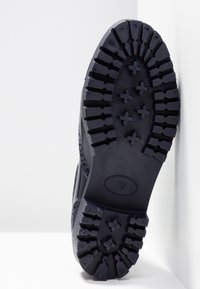 Semelle de chaussure noire avec des motifs de bande de roulement profonds et la taille 37 marquée au centre, positionnée contre des murs d'angle blancs.