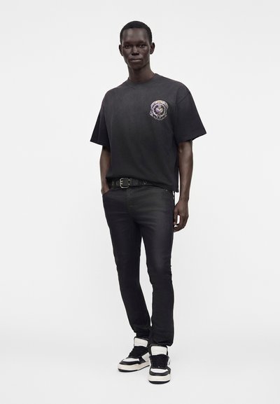 Άνδρας που φοράει μαύρο γραφικό t-shirt, μαύρο τζιν με ζώνη και μαύρα και λευκά ψηλά αθλητικά παπούτσια, στέκεται με το ένα χέρι στην τσέπη, σε λευκό φόντο.