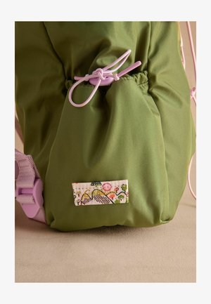 Grøn nylon taske med snørrelukning, lyserøde snore og en dekorativ label med et blomstermotiv og bjergmønster.