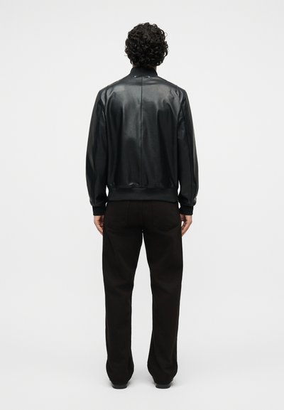 Άνδρας με σγουρά μαλλιά, φορούσε μαύρο δερμάτινο μπουφάν και μαύρο παντελόνι, στέκεται γυρισμένος με την πλάτη σε λευκό απλό φόντο.