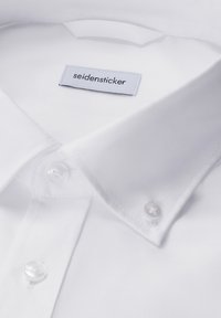 Biała koszula z eleganckim kołnierzykiem, zapinana na guziki, z naszywką z napisem "seidensticker" oraz gładką bawełnianą fakturą.