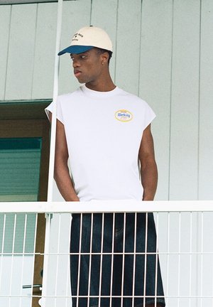 Jeune homme en débardeur blanc et casquette bicolore debout sur un balcon, regardant sur le côté, les mains dans les poches de son short foncé.