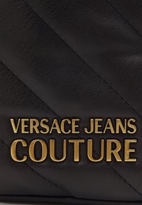 Černá kožená taška vyniká diagonálně prošívanou texturou s zlatými kovovými detaily, na které je výrazně zobrazeno "VERSACE JEANS COUTURE".