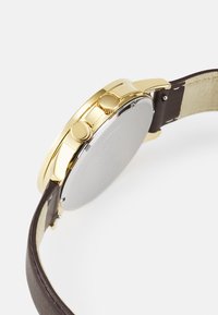 Armbanduhr aus goldfarbenem Metall mit rundem Gehäuse, braunem Lederarmband und zwei Druckknöpfen. Die Rückseite ist silberfarben und glatt ohne Gravuren.