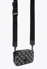 Zwarte geweven schoudertas met een glanzende afwerking, voorzien van een afneembare schouderriem, ritssluiting en een logo-accent aan de voorkant.
