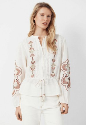 Jeune femme portant un chemisier blanc avec une broderie florale marron et verte et une taille nouée, assorti à un pantalon blanc sur un fond clair.